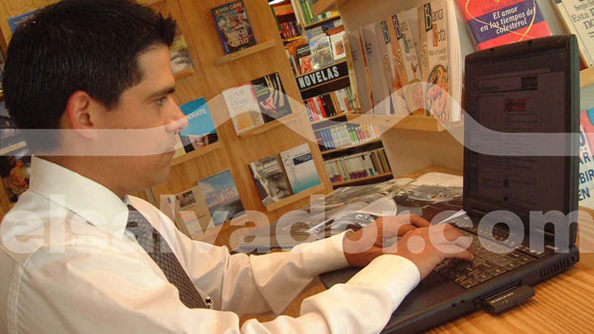 Hombre con camisa formal y corbata navegando en una computadora portátil antigua dentro de una librería rodeado de estantes de libros.