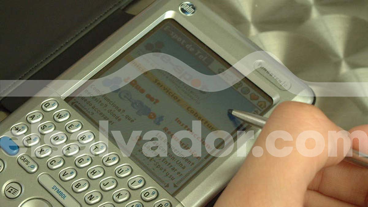 Mano sosteniendo un stylus para navegar en una PDA Palm Tungsten con teclado FSK mostrando la plataforma e-spot de Telecom El Salvador.