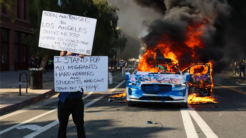 Un activista sostiene carteles mientras un coche arde durante las protestas provocadas por las redadas migratorias en Los Ángeles, California, EE. UU., el 8 de junio de 2025. El presidente estadounidense, Donald Trump, ha desplegado 2.000 efectivos de la Guardia Nacional, a pesar de no haber recibido ninguna solicitud de asistencia adicional del estado de California, tras las multitudinarias protestas contra las continuas redadas migratorias en el área de Los Ángeles durante los últimos días. (Protestas) EFE/EPA/ALLISON DINNER