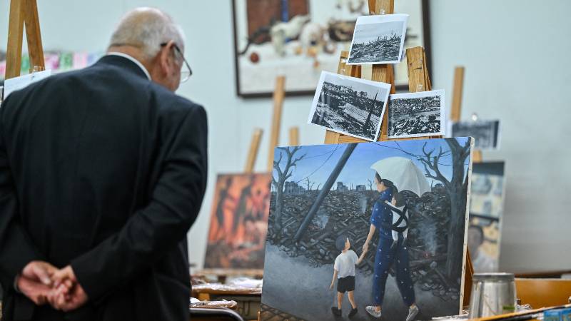 Esta foto, tomada el 29 de mayo de 2025, muestra a Masaki Hironaka, superviviente de la bomba atómica, observando una pintura (C) creada por la estudiante Hana Takasago, que lo representa caminando con su madre tras la devastación de 1945. / 