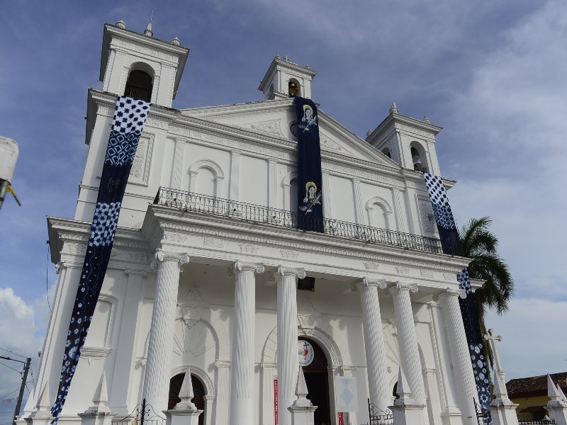 "La imponente fachada de la parroquia Santa Lucía, con sus seis columnas jónicas y su torre campanario, domina el centro histórico de Suchitoto."