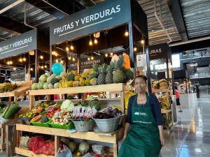 Vendedora en puesto de frutas y verduras del Mercado San Miguelito, rodeada de variedad de productos frescos como piñas, melones, tomates y hortalizas.