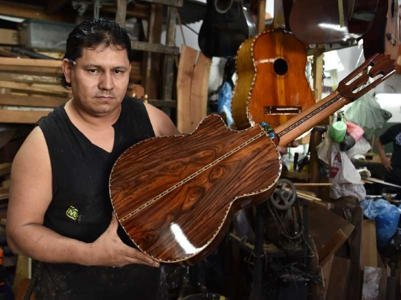 En el taller cuelgan guitarras, guitarrones y violines en distintas etapas de ensamblaje, creando un ambiente sonoro y artesanal.
