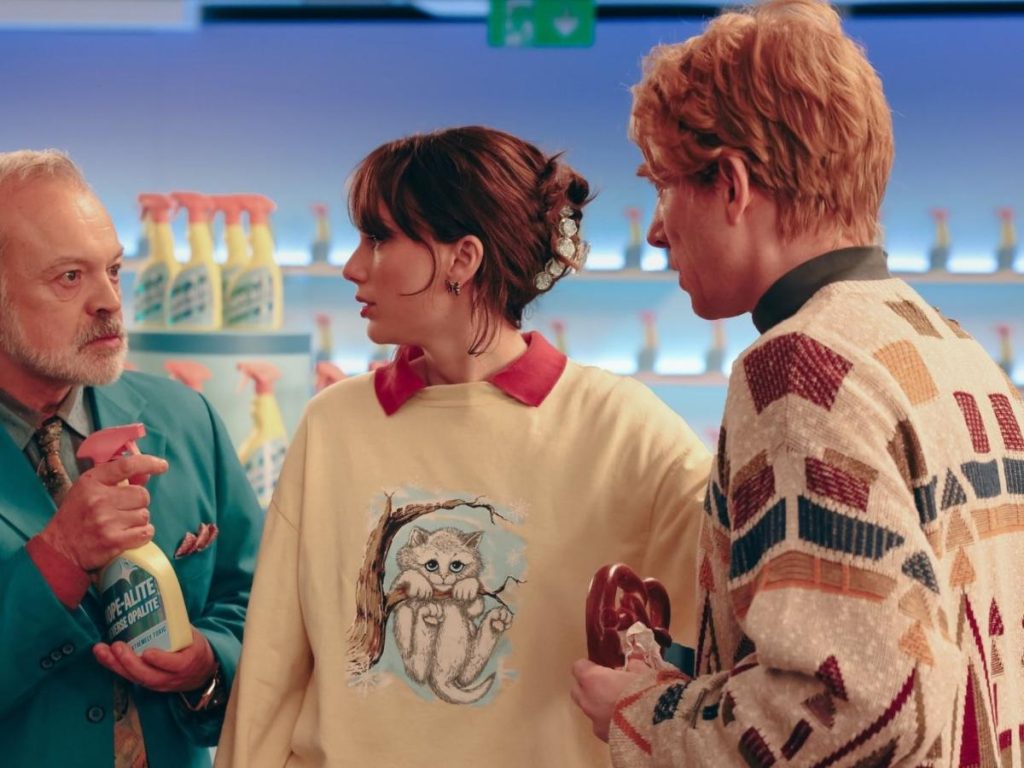 Una escena cargada de humor y estética retro, donde la protagonista interactúa con personajes secundarios en un entorno que recuerda a anuncios domésticos noventeros.