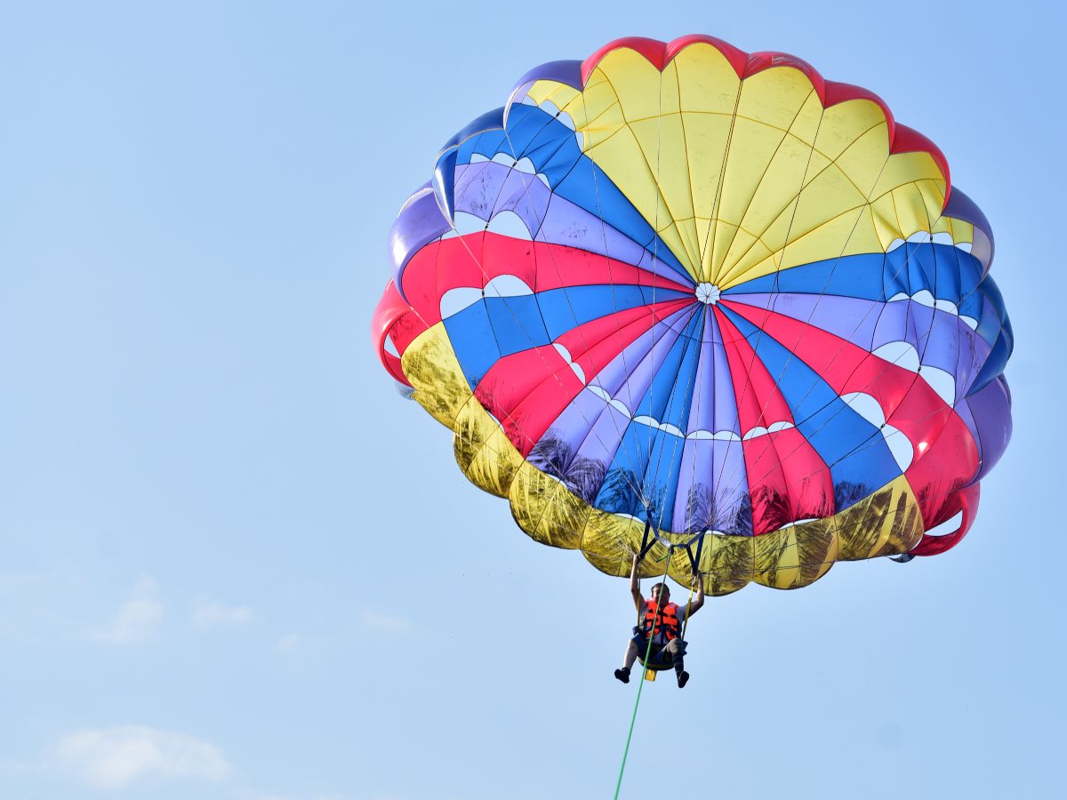 Durante el vuelo, el participante se eleva sostenido por un arnés y un paracaídas multicolor, mientras la lancha mantiene la tensión de la cuerda sobre el mar.