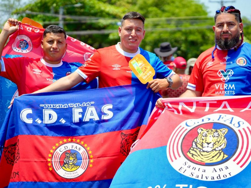 Un grupo de jóvenes aficionados de FAS ondea enormes banderas rojo y azul en las afueras del estadio, mientras uno de ellos destaca con trenzas adornadas con listones de los colores del equipo. Foto: cortesía La Primera