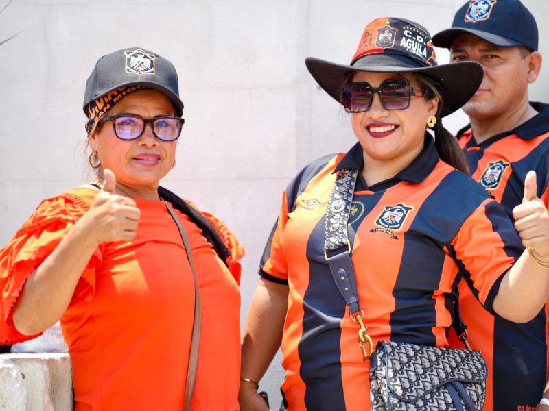 Dos aficionadas del Águila lucen orgullosas sus sombreros y camisetas en negro y anaranjado durante la fiesta futbolera del clásico nacional. Foto: cortesía Club Deportivo Aguila