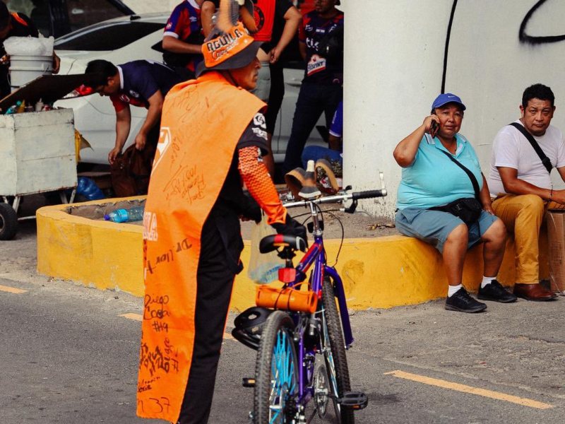 Un aficionado del Águila recorre los alrededores del estadio en bicicleta, luciendo una llamativa capa anaranjada para demostrar su pasión por el equipo emplumado. Foto: cortesía Club Deportivo Aguila