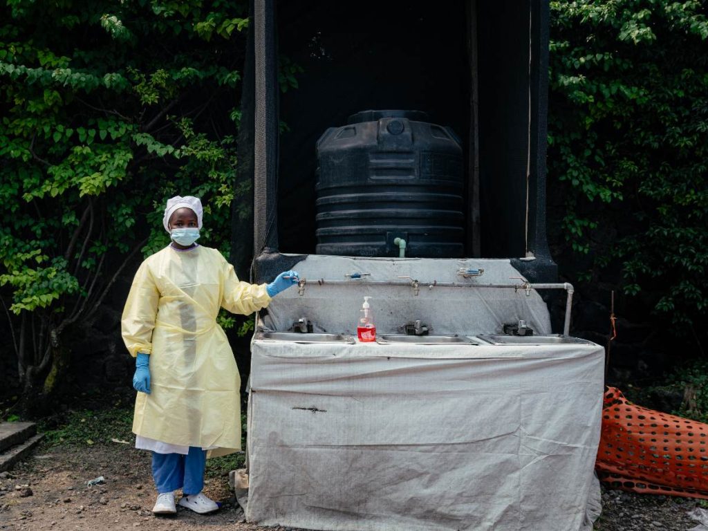 Una trabajadora sanitaria, vestida con ropa protectora desechable y gorro quirúrgico, se encuentra en un puesto de control donde supervisa el lavado de manos y la toma de temperatura a todos los visitantes y pacientes que ingresan al Hospital Kyeshero, como parte de las medidas de prevención del Ébola en Goma el 18 de mayo de 2026.