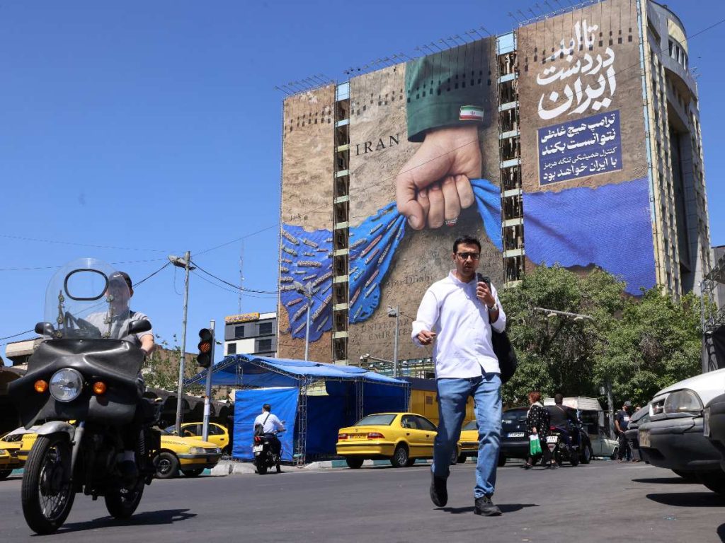 Varias personas cruzan una calle junto a una valla publicitaria en la fachada de un edificio que representa el estrecho de Ormuz con un letrero en persa que dice "Siempre en manos de Irán", en la plaza Vanak de Teherán, el 25 de mayo de 2026.