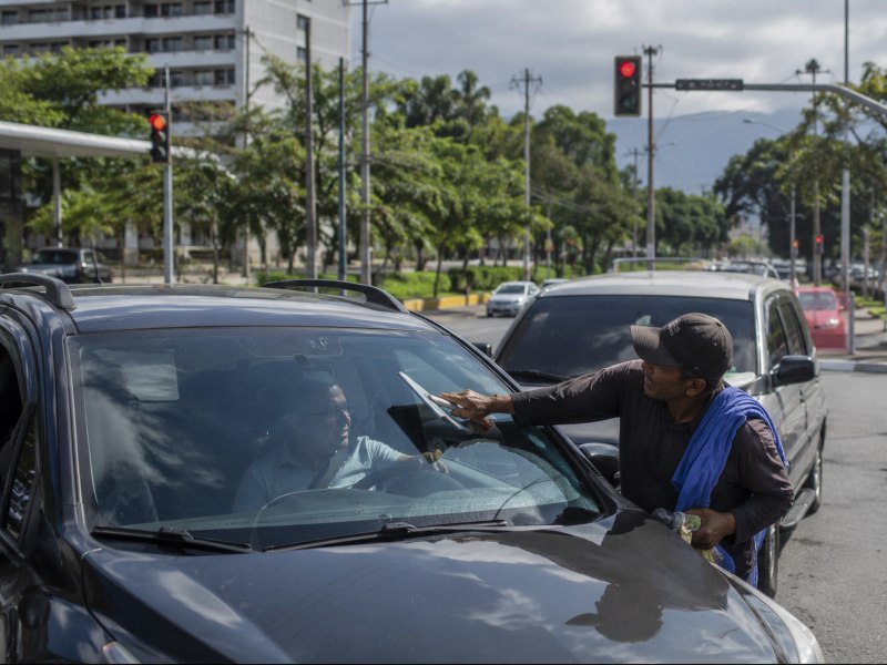 “Mientras el semáforo permanece en rojo, un hombre ofrece limpieza de parabrisas a conductores capitalinos.”