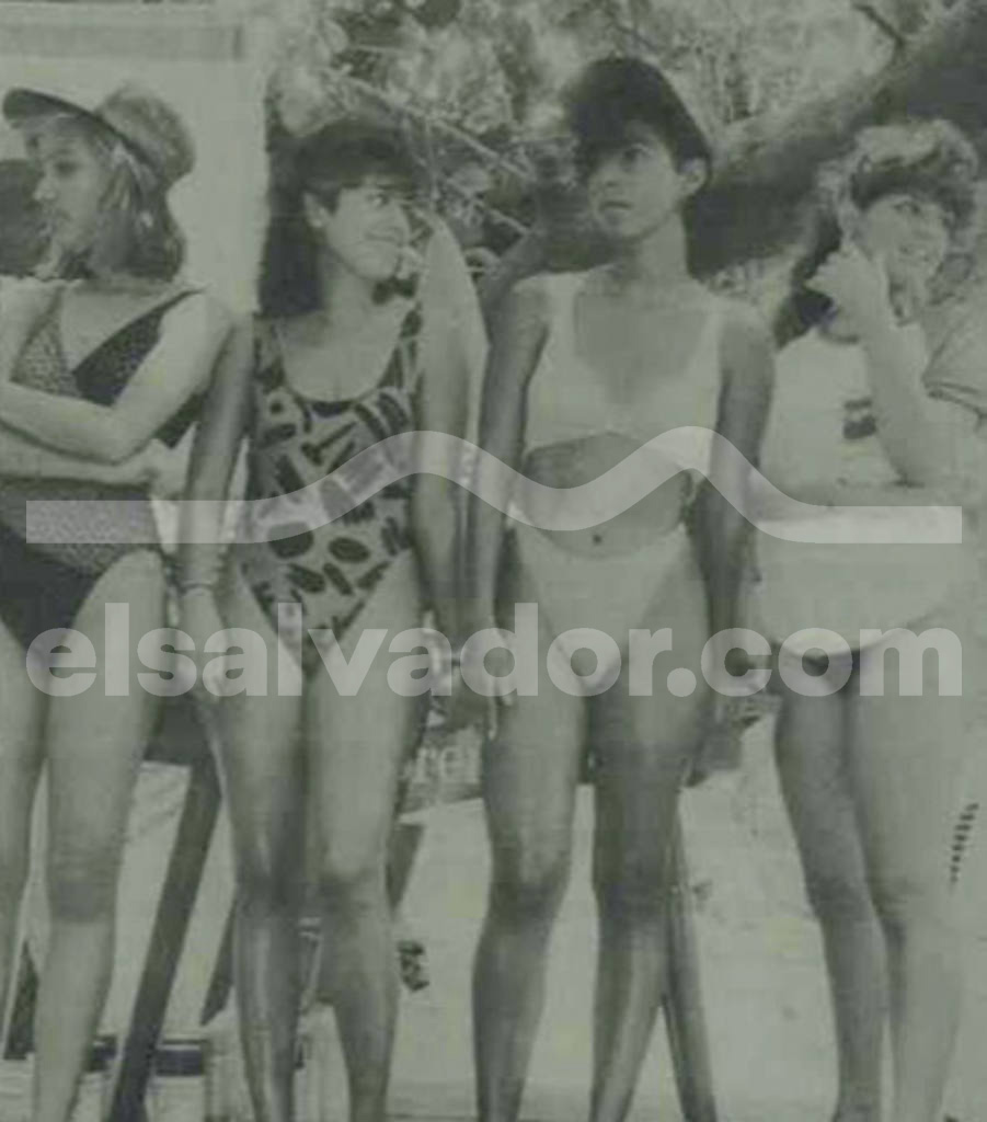 Fotografía antigua y difusa en blanco y negro de cuatro jóvenes salvadoreñas posando juntas en la playa a finales de los años 80. Llucen diferentes trajes de baño, incluyendo un bikini claro de dos piezas y un traje entero con estampado geométrico oscuro. Tienen cortes de cabello voluminosos propios de la época.