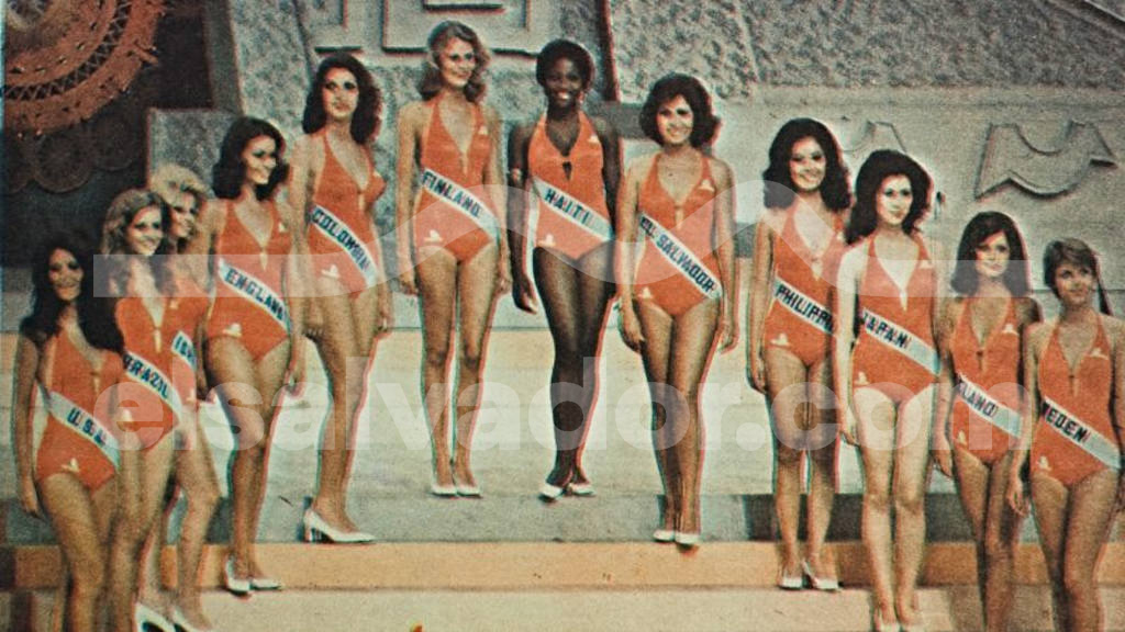 Fotografía a color de archivo histórico que muestra a un grupo de unas doce candidatas de Miss Universo 1975 posando alineadas sobre un escenario en San Salvador. Todas visten trajes de baño enteros idénticos de color naranja con bandas blancas de sus respectivos países, entre ellas Miss El Salvador, Miss USA y Miss Japón.