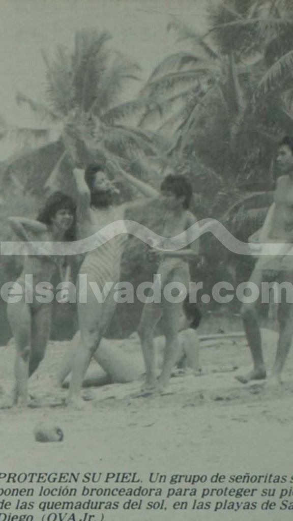 Fotografía antigua en blanco y negro con grano fotográfico grueso. Muestra a un grupo de jóvenes, hombres y mujeres en trajes de baño y bikinis, compartiendo en una playa tropical rodeada de palmeras en San Diego, La Libertad. Dos de las mujeres simulan aplicarse bronceador en los brazos.