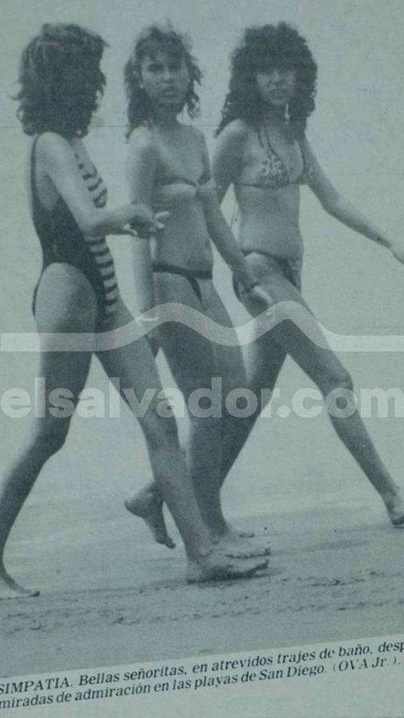Fotografía en blanco y negro tomada de perfil de tres mujeres jóvenes con peinados esponjosos de los años 80 caminando descalzas sobre la arena húmeda de la playa. La primera viste un traje entero oscuro con rayas blancas en los costados y corte alto, y las otras dos llevan bikinis de dos piezas.