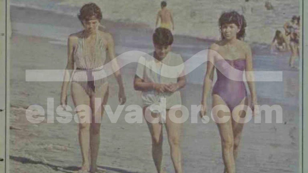 Fotografía a color con tonos desgastados que muestra a tres personas caminando de frente por la orilla del mar en una playa con oleaje suave. A la izquierda, una mujer con traje entero a rayas finas; al centro, un joven con camiseta clara; a la derecha, una mujer con un traje de baño entero color violeta.