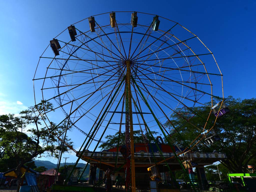 Trabajadores ensamblan la enorme estructura de metal de la Rueda de Chicago en el Campo de la Feria de Santa Ana. Al fondo, se aprecian frondosos árboles y montañas bajo un cielo azul claro.