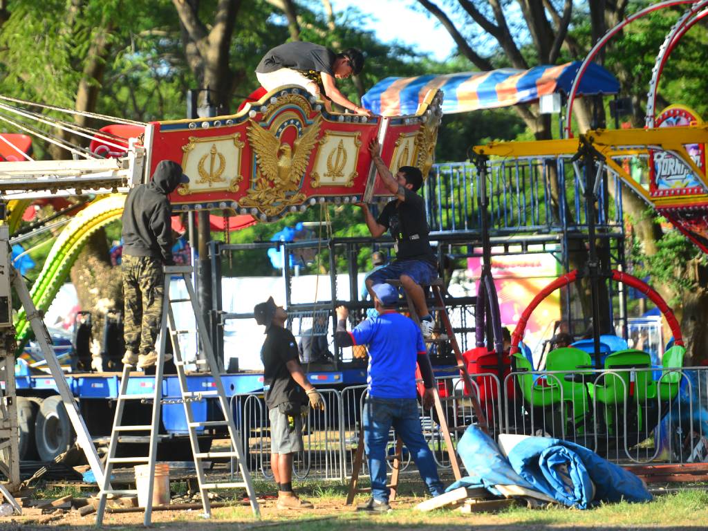 Fotografía en plano medio que muestra a un equipo de trabajadores en el campo de la feria instalando la corona decorativa de un carrusel. Dos operarios subidos en escaleras sostienen un gran panel rojo y dorado ornamentado con el relieve de un águila, mientras un compañero trabaja en la parte superior y otros dos supervisan la instalación desde el suelo. Al fondo se aprecian árboles y otras atracciones mecánicas en preparación.