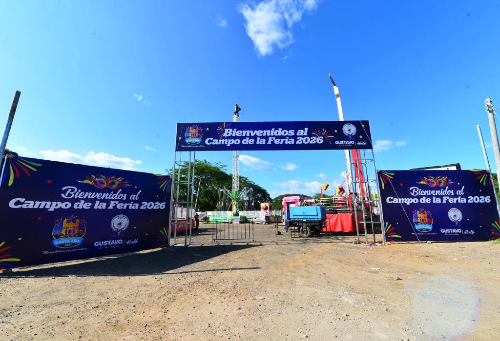 Fotografía que muestra la entrada principal del Campo de la Feria. Destaca una gran estructura de bienvenida en forma de arco decorada con motivos festivos y coloridos, junto a senderos limpios y recién acondicionados. Al fondo se alcanza a ver el terreno de la feria en preparación, con ruedas mecánicas en proceso de instalación bajo un cielo parcialmente despejado.