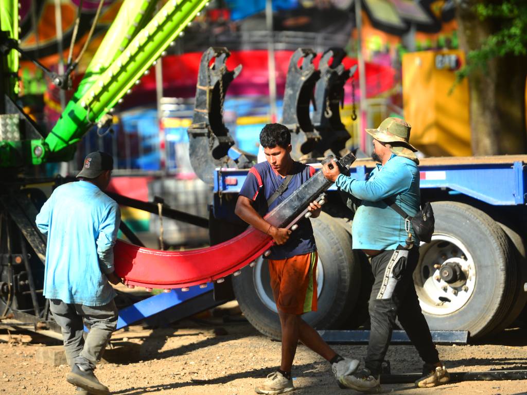 Un grupo de operarios y técnicos en un campo ferial trabaja en equipo para levantar y sostener una gran pieza curva de hierro, correspondiente a uno de los tentáculos del juego mecánico "El Pulpo". Al
