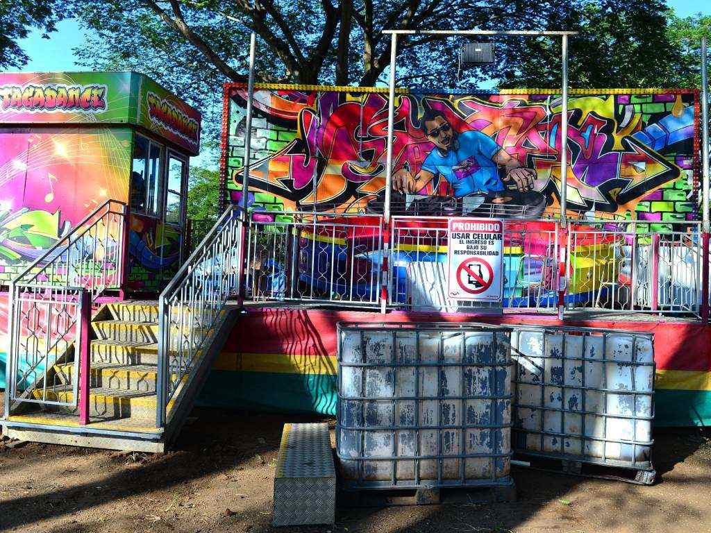 Fotografía de la atracción de feria "Tagadance", una versión del clásico Tagadá, completamente armada en un terreno al aire libre. Al fondo resalta un mural estilo grafiti de colores vibrantes con la ilustración de un DJ, junto a la cabina de control a la izquierda. En el centro de la barandilla metálica de acceso se observa un cartel de advertencia que prohíbe el uso de teléfonos celulares dentro del juego mecánico.