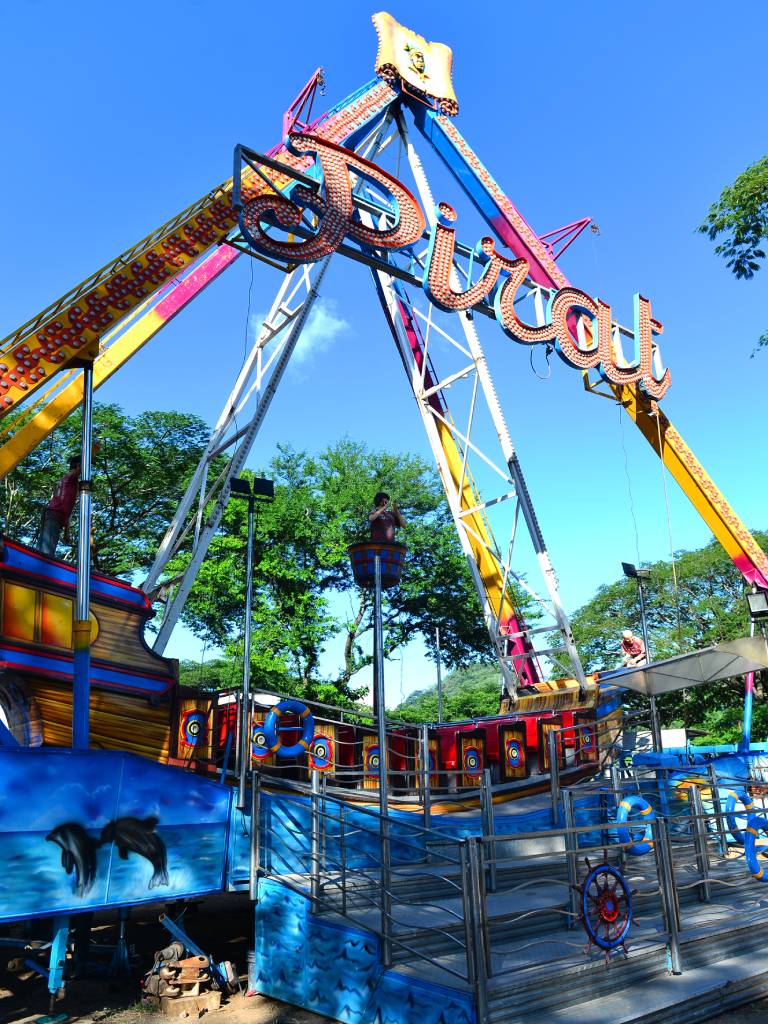 Ángulo contrapicado de la atracción mecánica "PIRAT" (Barco Pirata) en el campo de la feria. Se observa la gran estructura metálica de soporte pintada de amarillo y blanco con un gran rótulo luminoso en el arco. En la base, el barco imita un galeón antiguo de madera, rodeado por barandillas metálicas decoradas con timones y salvavidas, mientras varios técnicos realizan labores de mantenimiento previo a su inauguración.
