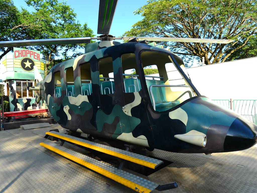 Fotografía en primer plano de una cabina infantil de feria con forma detallada de helicóptero militar, pintada con camuflaje en tonos verde, beige y negro. La estructura, llamada "CHOPPER", ya se encuentra armada sobre su plataforma, mostrando sus ventanas abiertas y asientos internos listos para recibir a los niños.