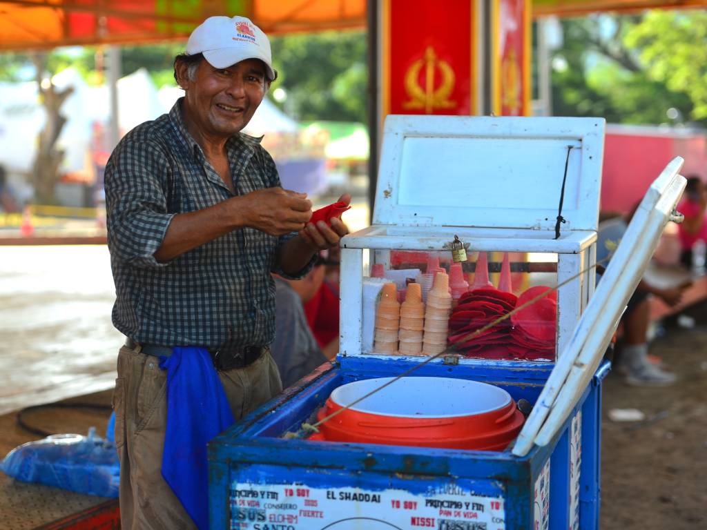 amuel Durán, un vendedor de 58 años, sonriendo amablemente junto a su tradicional carretón de sorbete artesanal en el campo de la feria. El carretón está decorado con colores vivos y llamativos, y en la parte superior se observan los barquillos y recipientes metálicos listos para atender a los visitantes de las festividades.