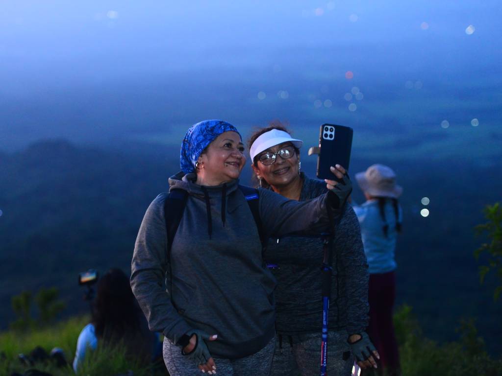 ENFOQUE EN CONQUISTAR CADA RUMBO. Fotografía en plano amplio que muestra a varias senderistas de Rumbo Femenino caminando en fila o alcanzando juntas la cima de un terreno volcánico, rodeadas de nubes y un cielo despejado.