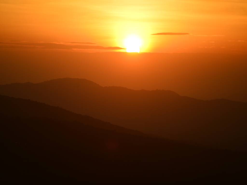 DESTINO: CERRO EL CHINO. Fotografía de un majestuoso amanecer desde la cima del Cerro El Chino. El sol brilla intensamente en un cielo teñido de tonos dorados y anaranjados, mientras que en la parte inferior se aprecian las siluetas oscuras de las montañas en contraluz.