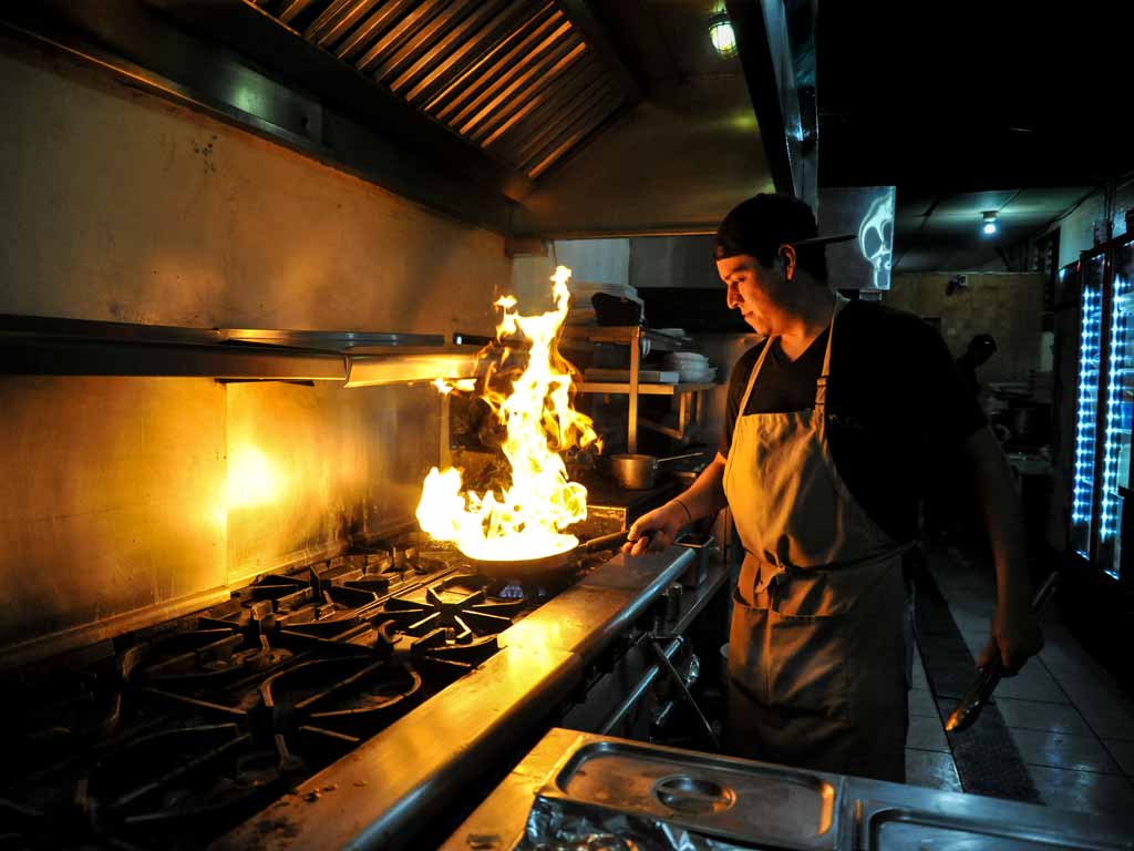 Un cocinero prepara comida enfrente de la cocina con una olla en llamas en Inna Jammin Santa Ana.