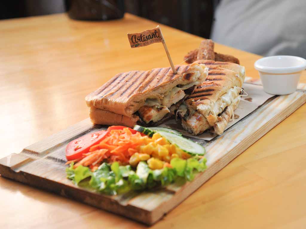 Un plano de detalle elevado que muestra una porción de un panini a la parrilla relleno de pollo, queso derretido y champiñones, presentado sobre una tabla de servir de madera texturizada en una mesa de madera.