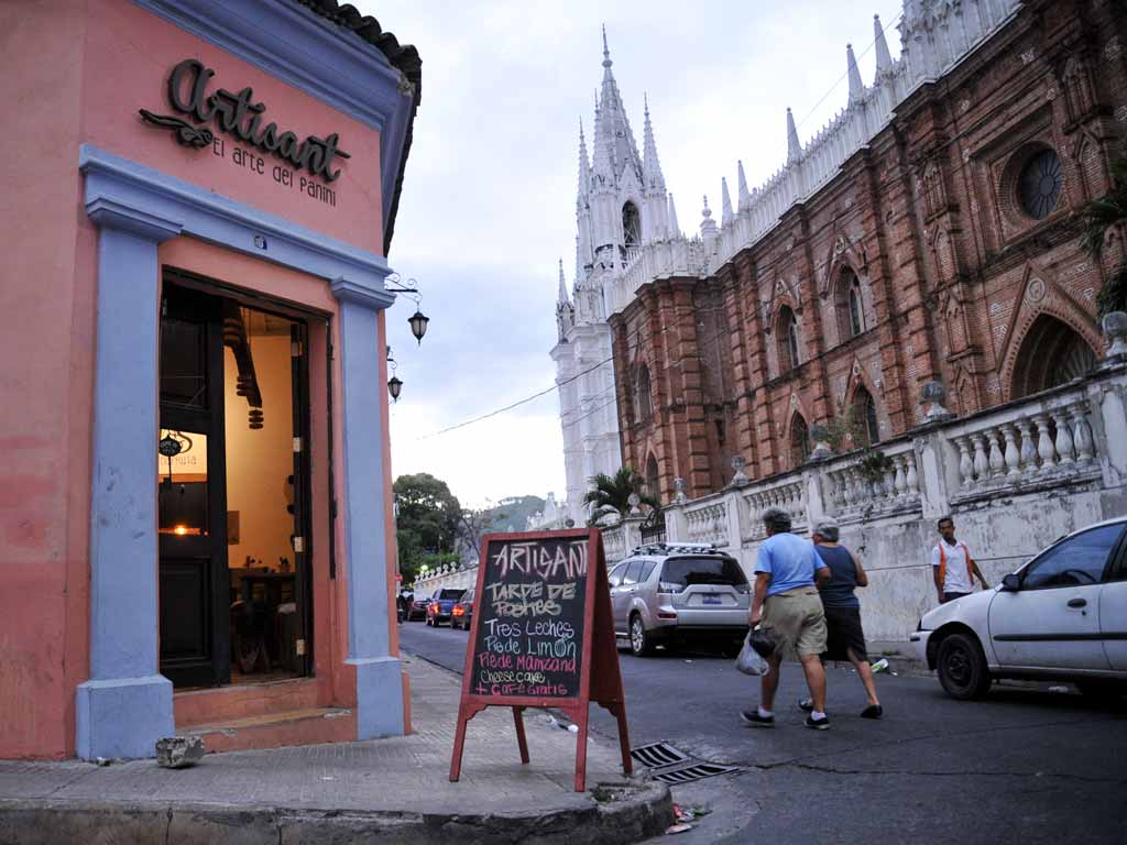 Una toma de esquina de la colorida fachada del restaurante Artisant en Santa Ana. La pared de estuco rosa tiene un letrero negro con la frase "Artisant, El arte del panini" y una puerta de madera abierta con luces cálidas en su interior.