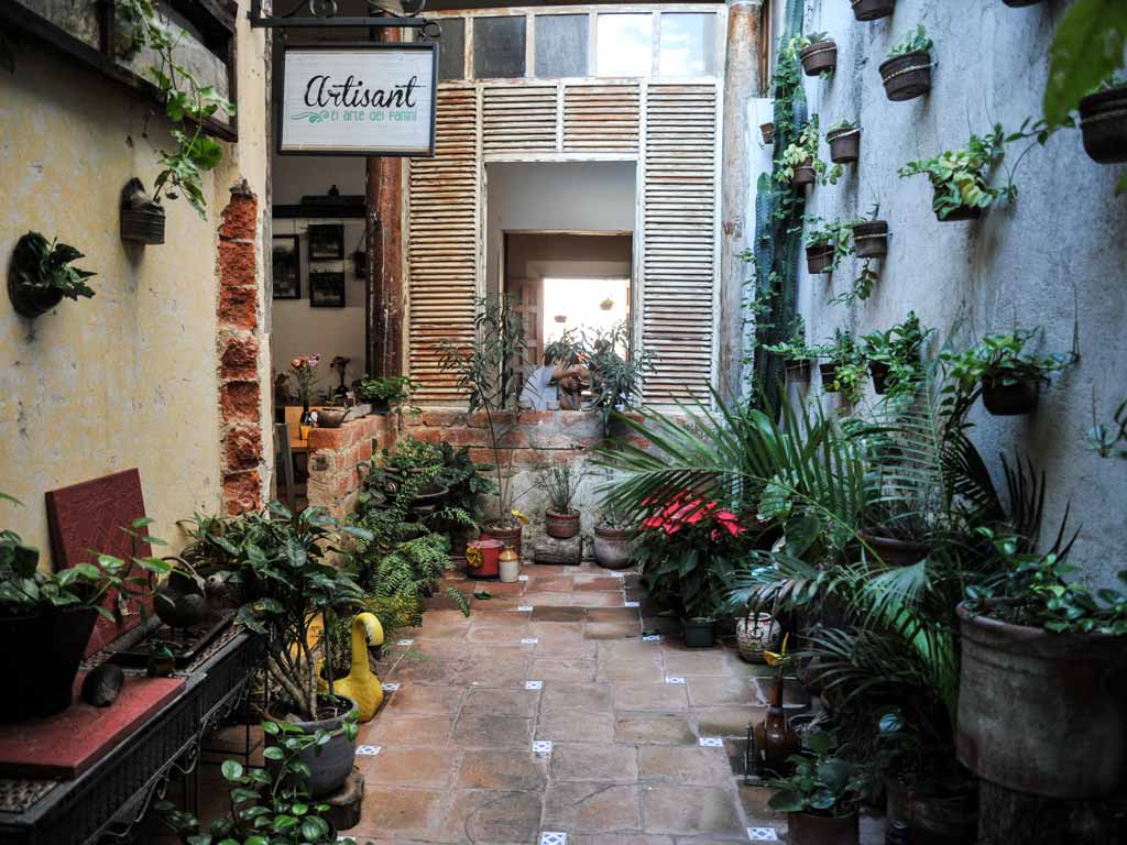 Una vista que mira a través de un pasillo al aire libre lleno de plantas en macetas en Artisant. El pasillo de azulejos rústicos está bordeado por paredes de estuco amarillo envejecido y pared de ladrillo visto a la izquierda y una pared texturizada llena de macetas de plantas colgantes a la derecha.