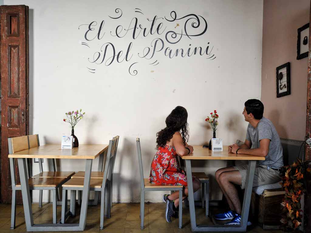 El interior de un café rústico y acogedor en Artisant. Una pareja joven, una mujer con un vestido floral rojo y un hombre con una camiseta gris, están sentados en una mesa de madera clara con patas de metal industrial, sonriendo y conversando.
