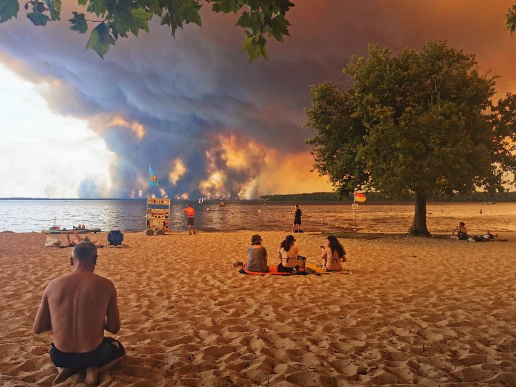 Bañistas descansan en la playa de Moutchic, mientras una columna de humo del incendio en Gironda se eleva al fondo. Fotografía/ AFP