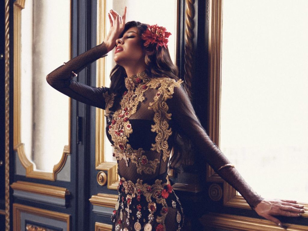 La reina lució un vestido negro con transparencias, bordados dorados y flores rojas, complementado con maquillaje intenso y accesorios dorados.