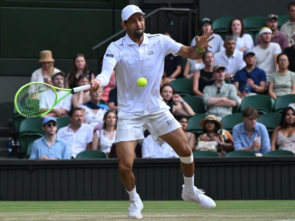 Una toma de acción en primer plano de Marcelo Arévalo en plena cancha de césped. Viste la indumentaria clásica e impecable de color blanco de Wimbledon (marca EA7) y una gorra a juego. Está totalmente enfocado, con los ojos fijos en la pelota de tenis que flota frente a él, mientras se prepara para conectar un golpe de derecha (forehand). Al fondo se puede apreciar al público en las gradas observando atentamente la jugada