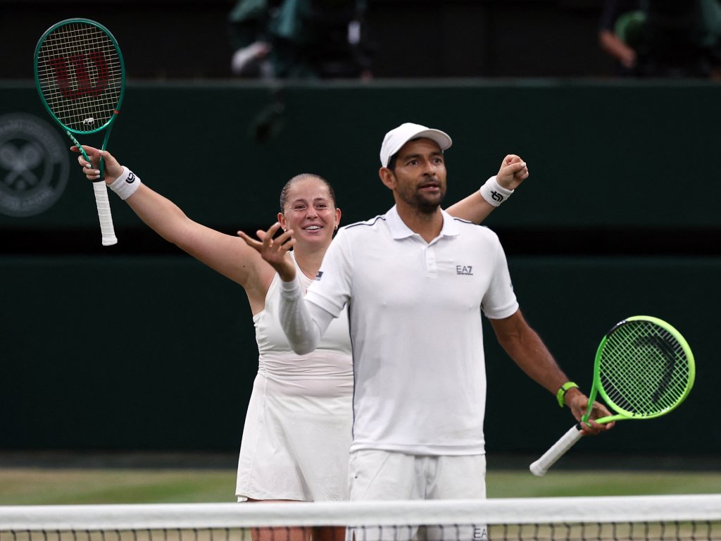 Una imagen llena de energía. Marcelo Arévalo celebra con euforia mirando hacia la grada o a su equipo, apretando el puño con fuerza y con el rostro reflejando pura adrenalina. A la izquierda, de espaldas a la cámara, Jelena Ostapenko camina hacia él levantando la mano en señal de festejo. El público en el fondo complementa la atmósfera de un momento crucial del partido.