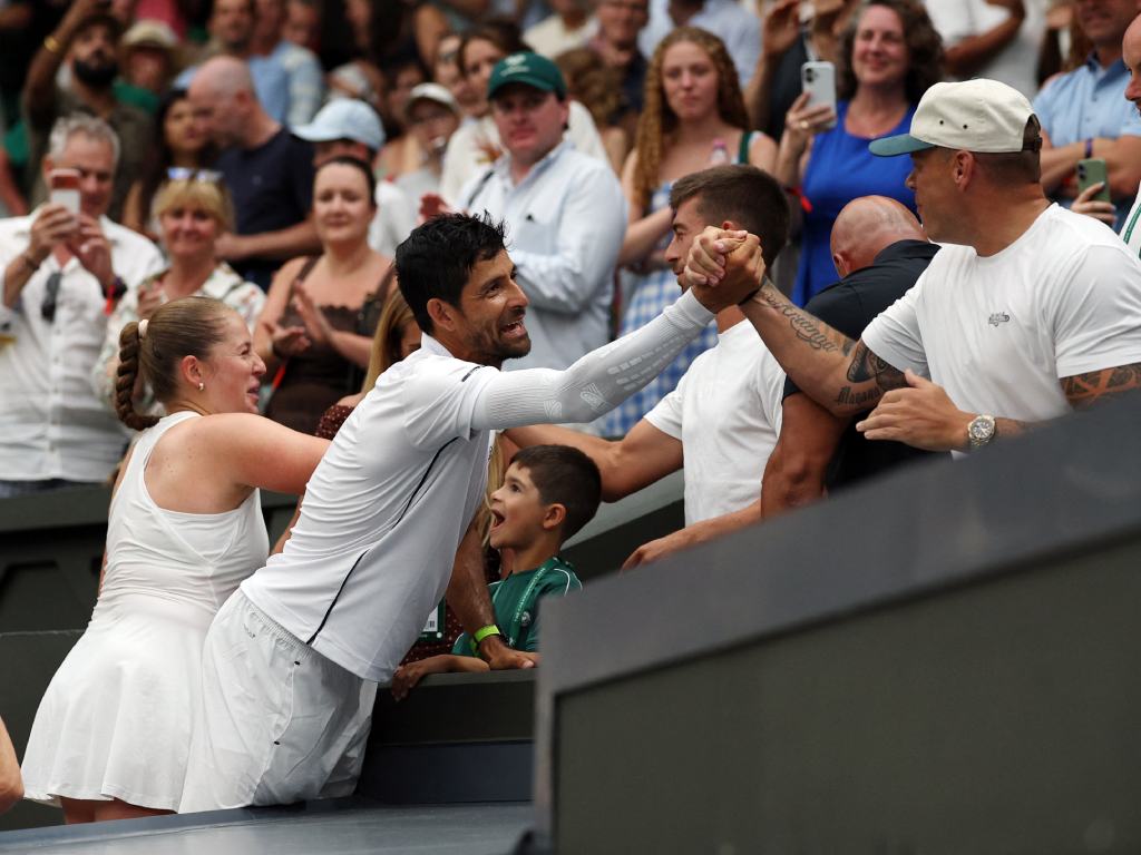 Un momento sumamente emotivo en el que Marcelo Arévalo se inclina sobre la baranda para estrechar fuertemente la mano de un miembro de su equipo o familiar en las gradas. A su lado, Jelena Ostapenko sonríe con alegría contagiada, mientras un niño pequeño en el público mira al tenista salvadoreño con una expresión de total asombro y felicidad.