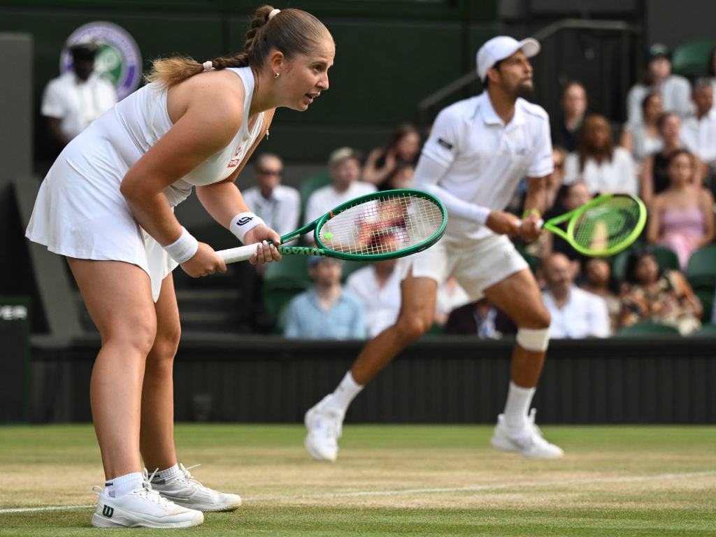 Una gran toma en perspectiva que muestra la sincronización de la pareja. En primer plano y enfocada, Jelena Ostapenko aparece semi-agachada, sosteniendo su raqueta verde con ambas manos, lista para recibir el servicio rival. En el fondo, un poco más rezagado pero en la misma postura de alerta, se observa a Marcelo Arévalo cubriendo la red. Ambos lucen el uniforme reglamentario blanco.