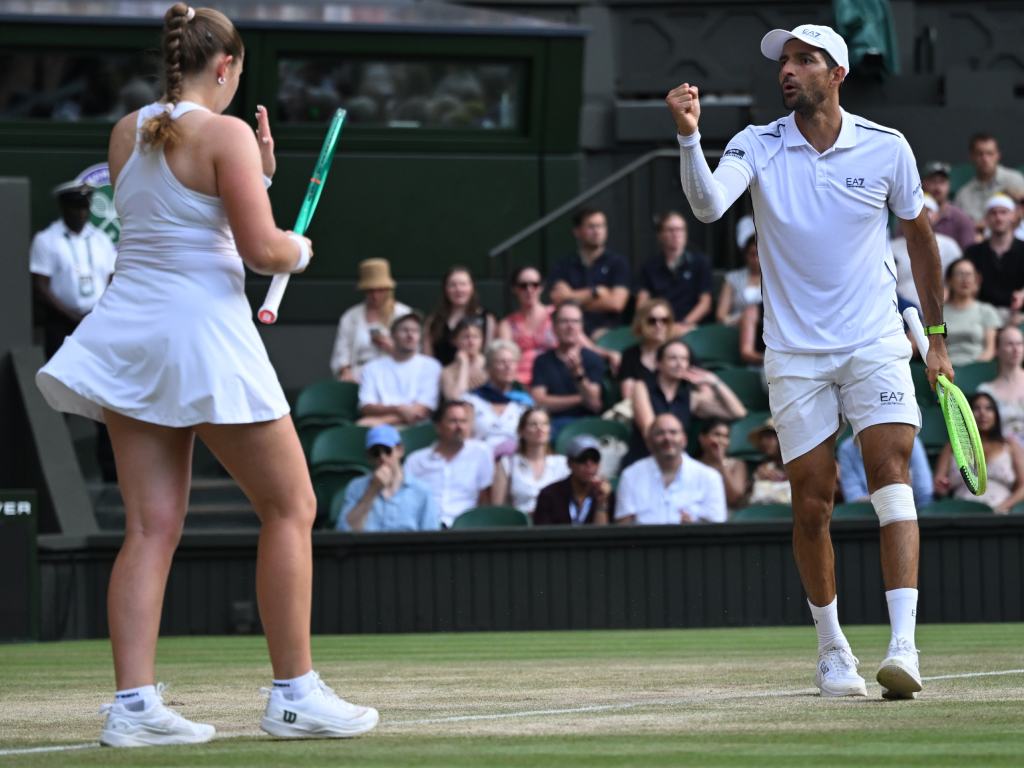 Justo después de sellar la victoria, la pareja celebra de cara al público. Jelena Ostapenko aparece detrás con los brazos abiertos y la raqueta en alto, mostrando una sonrisa radiante de oreja a oreja. Al frente, Marcelo Arévalo camina con el puño apretado y la mirada fija, absorbiendo los aplausos de las gradas de Wimbledon.