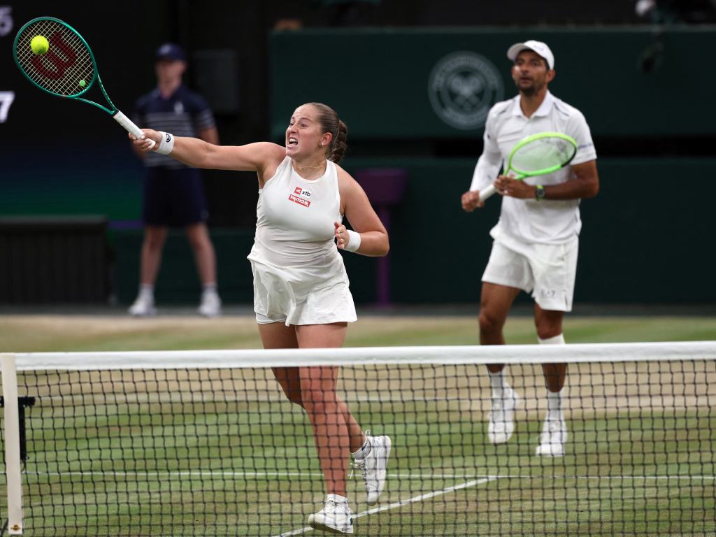 Captura dinámica en la que Jelena Ostapenko se estira con todo el cuerpo cerca de la red para impactar una volea exigida. Su rostro refleja el máximo esfuerzo físico y la intensidad del juego. Al fondo, Marcelo Arévalo sigue la jugada con la mirada, manteniéndose listo en caso de que la pelota regrese.