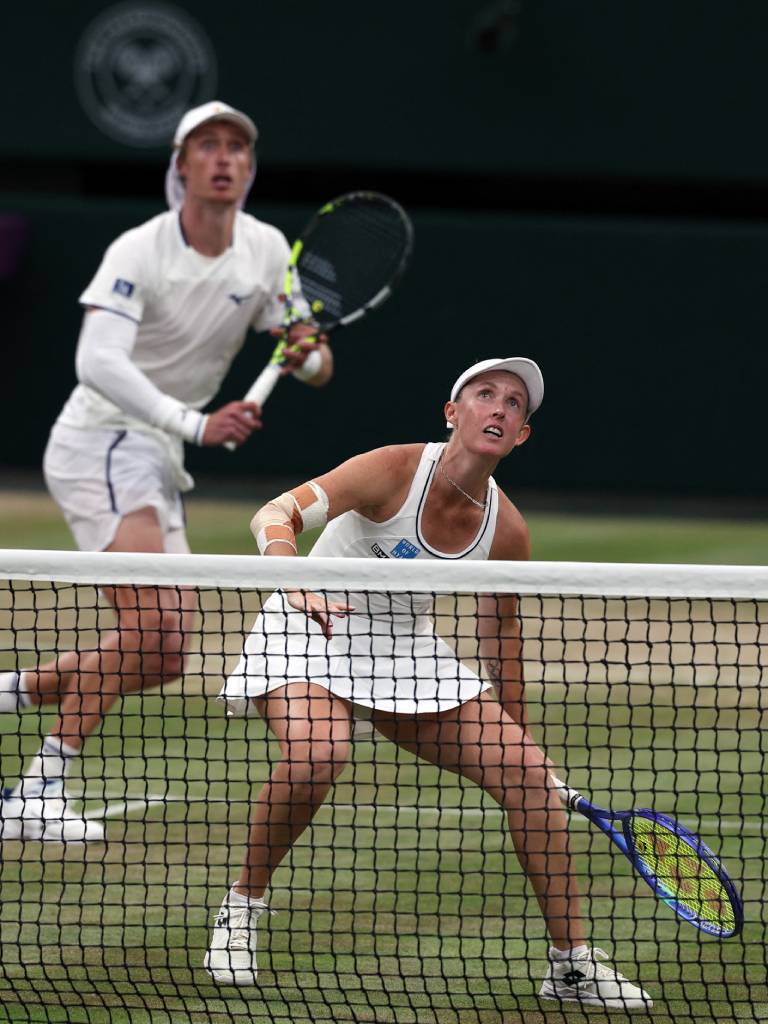 A diferencia de las anteriores, esta foto enfoca en primer plano a la pareja rival (otra dupla de dobles mixtos en Wimbledon) defendiendo la red. La jugadora en primer plano se encuentra agachada mirando hacia arriba, esperando un "globo" o un remate alto, mientras su compañero se posiciona detrás de ella con la raqueta en alto.