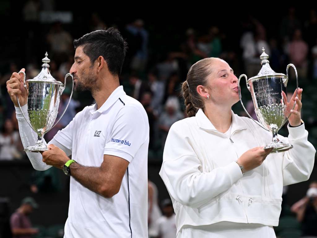 Una hermosa toma de perfil que captura a Arévalo y Ostapenko besando simultáneamente sus respectivos trofeos de campeones de Wimbledon. Los rostros de ambos reflejan una mezcla de alivio, orgullo y el tierno cumplimiento de un sueño deportivo en la catedral del tenis.