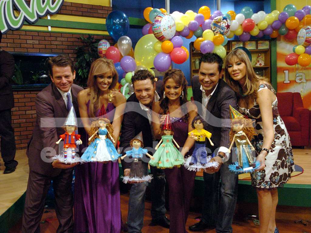 El elenco original posa sonriente rodeado de globos festivos mientras sostienen coloridas muñecas personalizadas hechas a mano, capturando un momento especial de aniversario en el set.