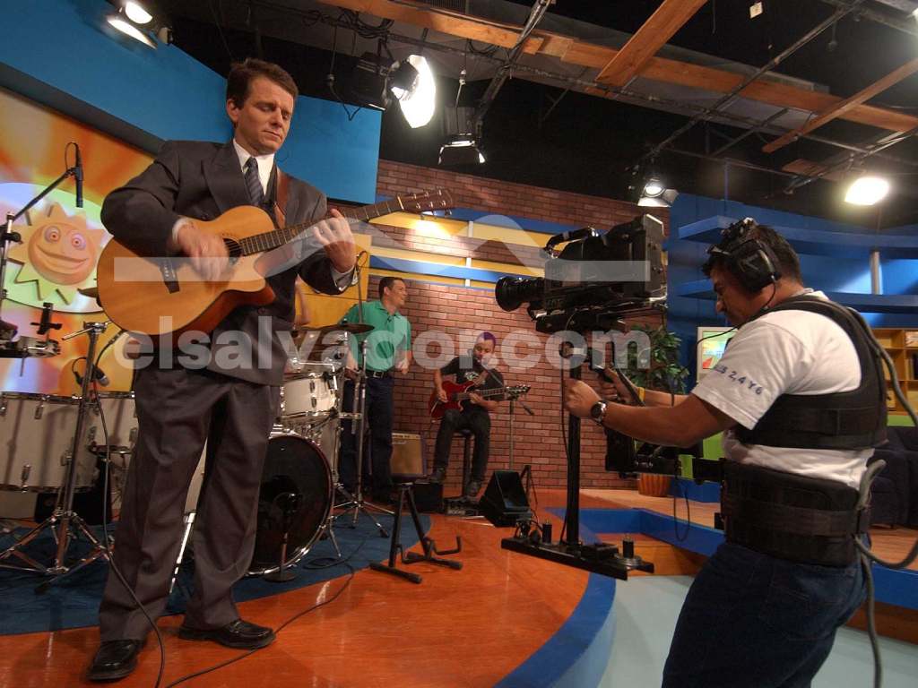 Un camarógrafo capta de cerca la interpretación en guitarra de uno de los presentadores en traje formal, apoyado por músicos en el fondo del set principal.