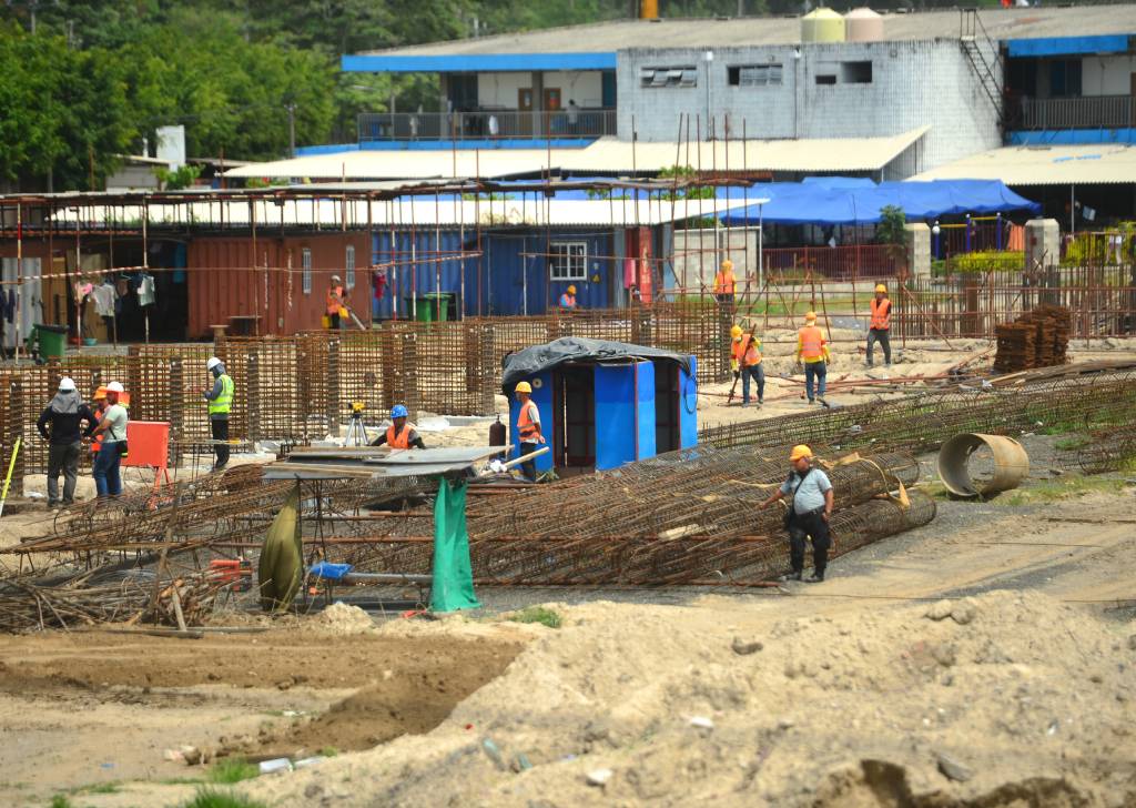 Grupo de obreros con cascos de seguridad realizan labores de construcción sobre el terreno y estructuras del estadio.