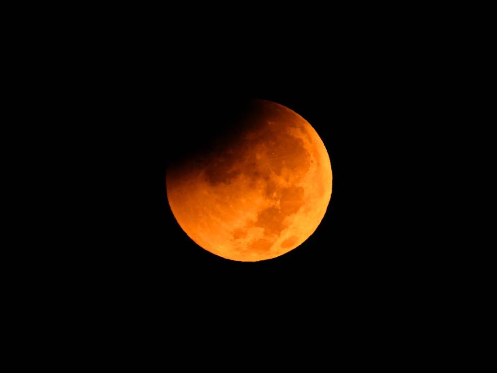 La Luna llena brillando en el cielo nocturno sobre el mar y los edificios iluminados de una ciudad costera.