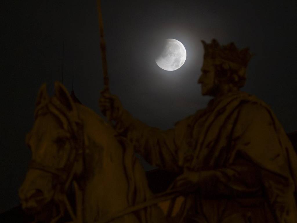 Silueta de una estatua ecuestre en primer plano con la Luna iniciando su eclipse en el cielo nocturno de una plaza.